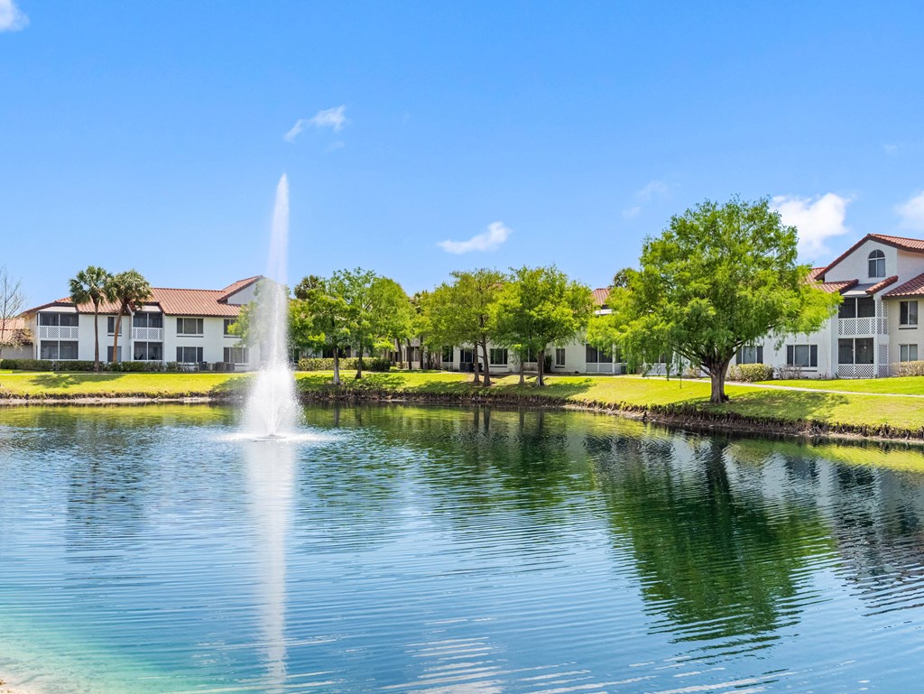 A fountain in the middle of a lake with houses in the background.